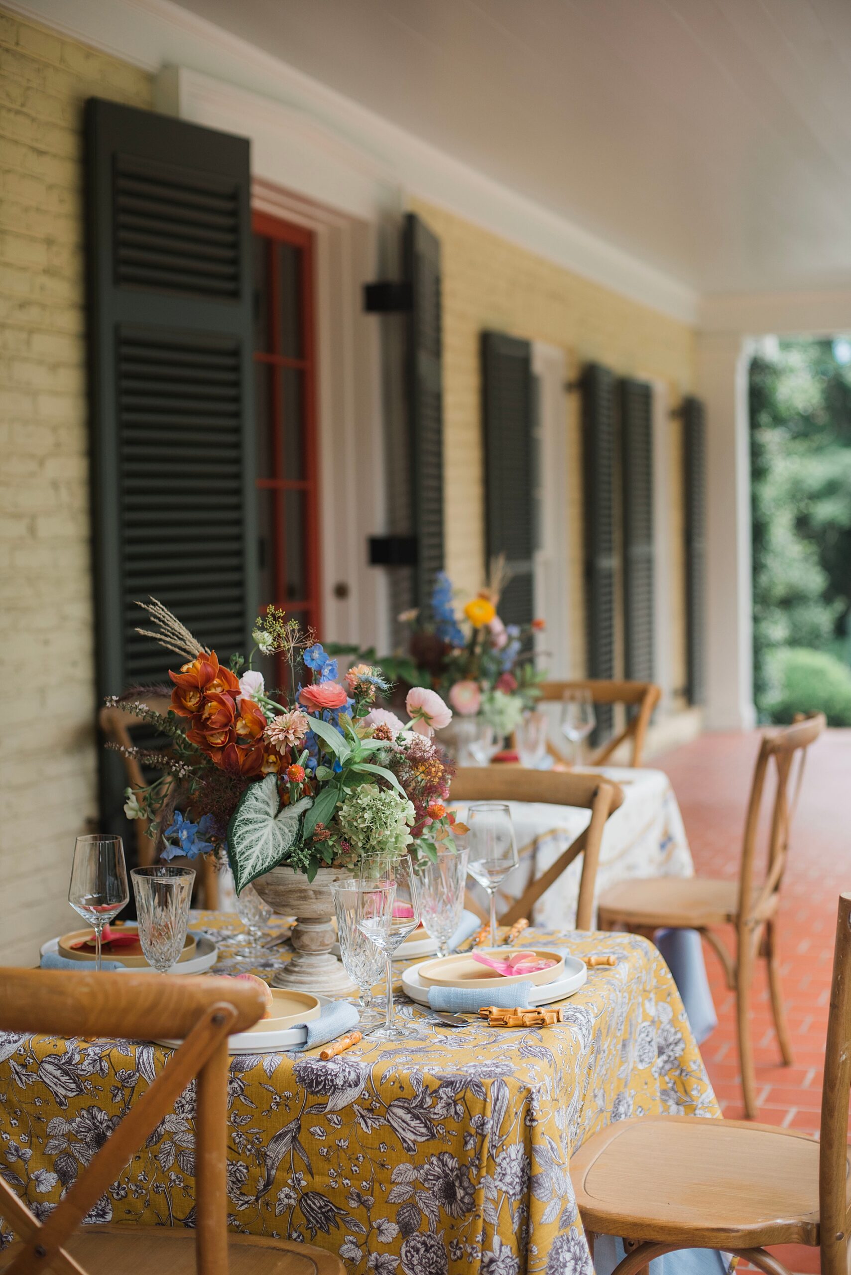 tables sit on covered porch decorated with flowers and ornate tablecloths.