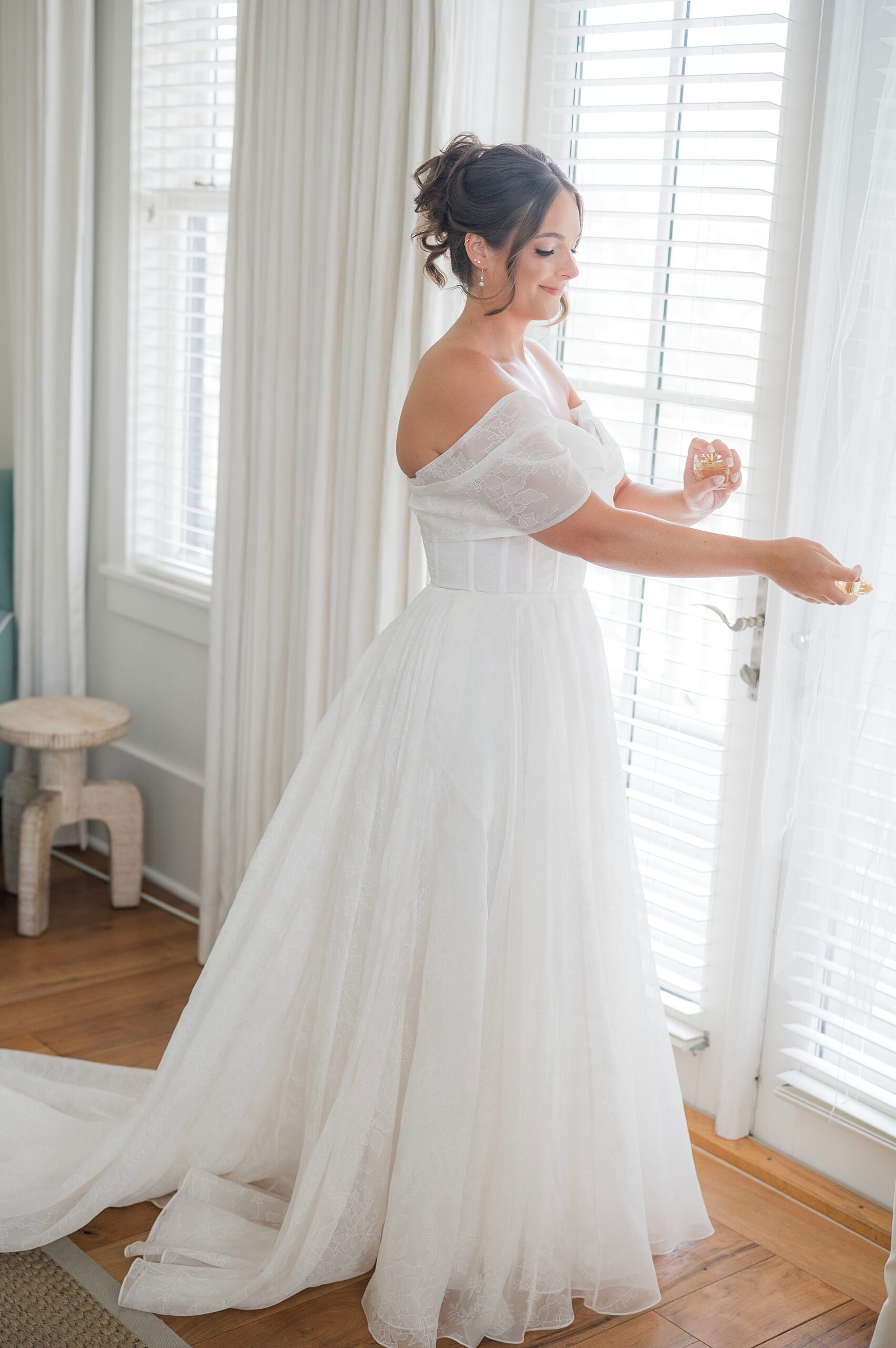bride stands by window and sprays perfume