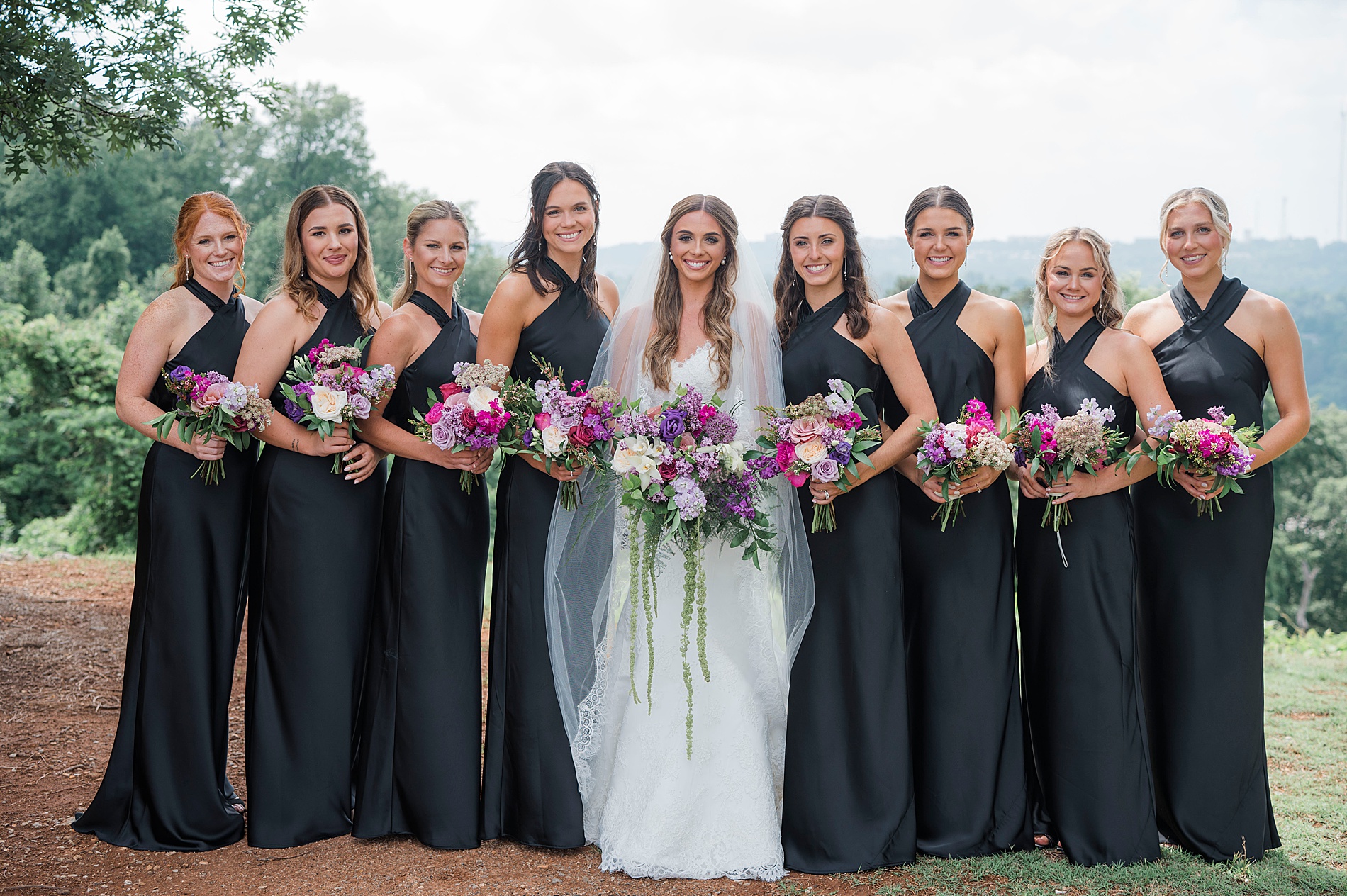 bridesmaids in black dresses with high neckline stand with bride all holding bouquets with white and purple flowers