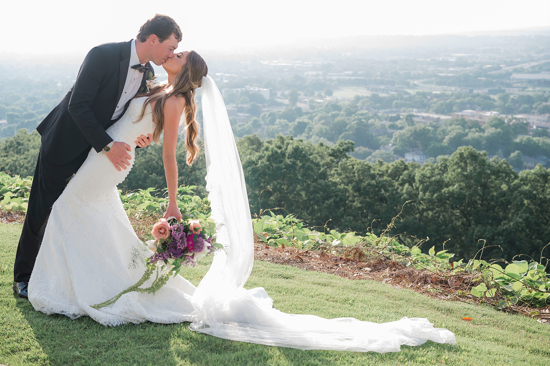 romantic wedding photos of groom kissing bride on hillside with a stunning view