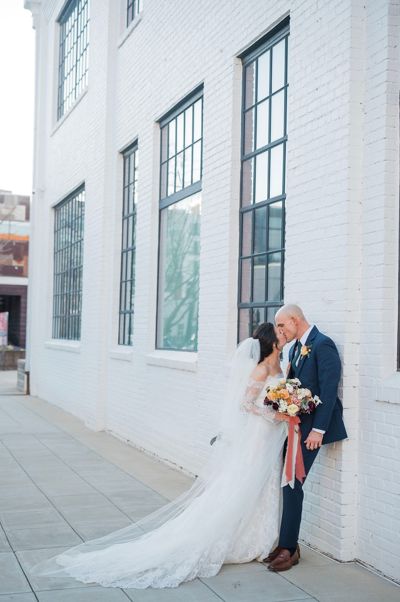 bride and groom kiss while leaning up against white brick wall